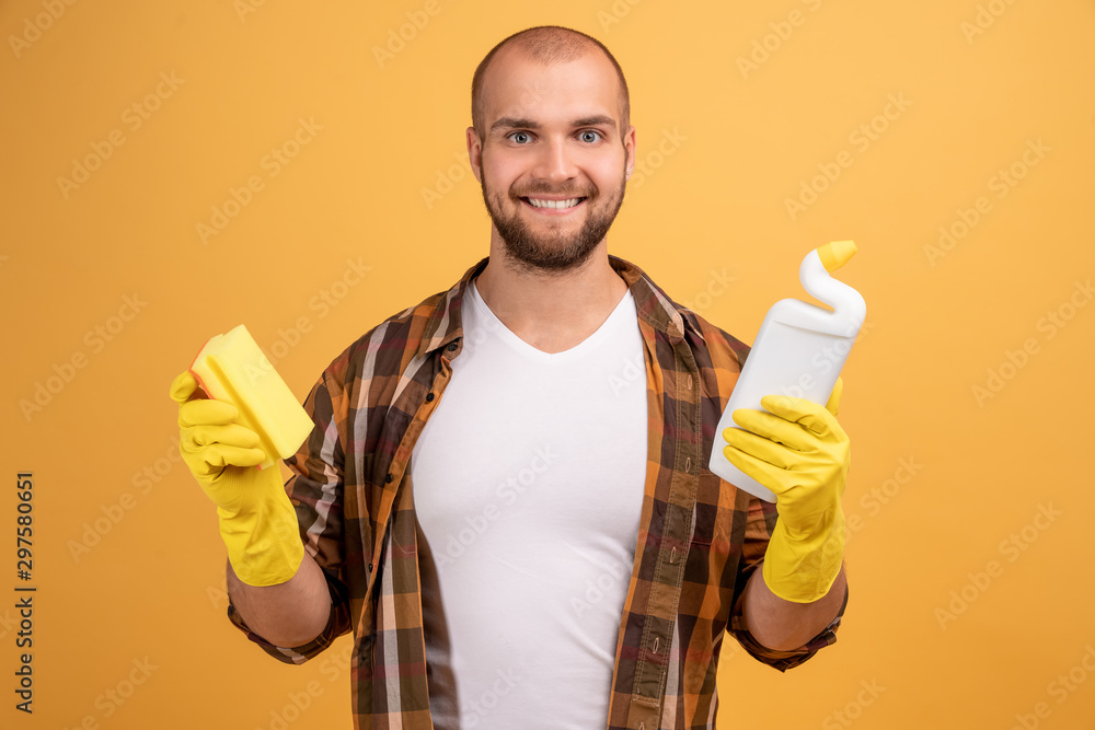 Happy bearded man dressed in casual clothes holding sponge and detergent going to clean house having good mood isolated over yellow background.Smiling cleaner with sponge and cleanser doing housework Happy bearded man dressed in casual clothes holding sponge and detergent going to clean house having good mood isolated over yellow background.Smiling cleaner with sponge and cleanser doing housework