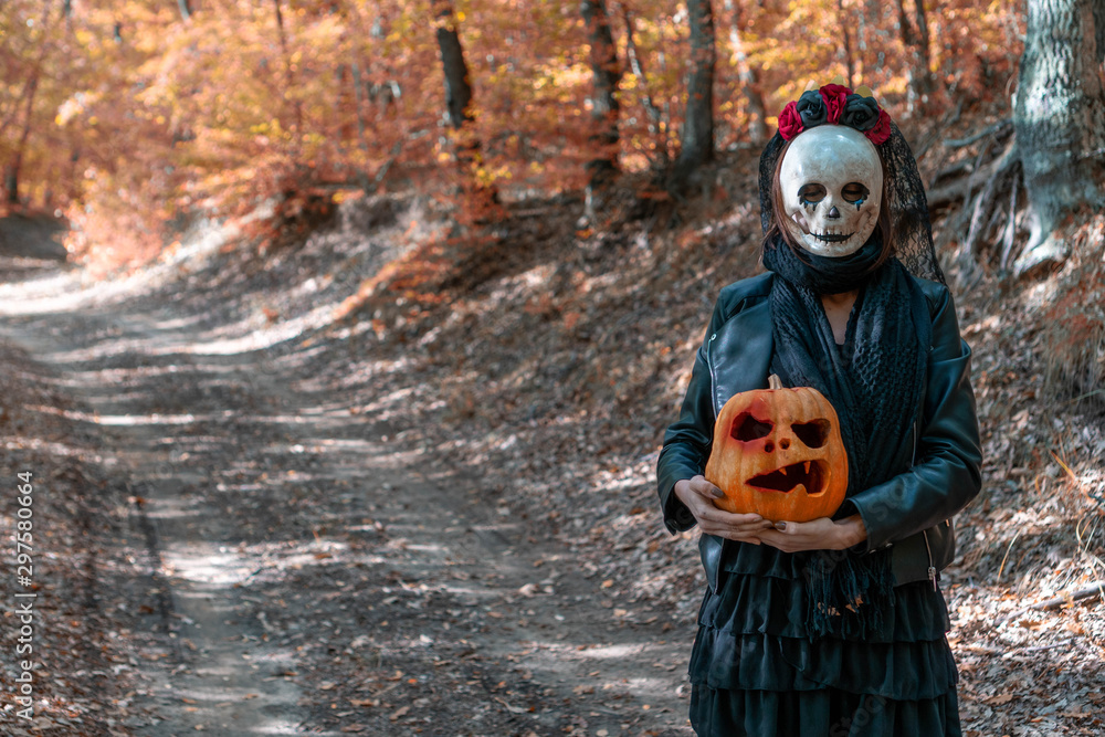 Photo of walking girl with creepy skull mask in the autumn woods, holding a carved Halloween pumpkin. On background path in autumn forest with fall leaves. Concept about Halloween. Photo of walking girl with creepy skull mask in the autumn woods, holding a carved Halloween pumpkin. On background path in autumn forest with fall leaves. Concept about Halloween.