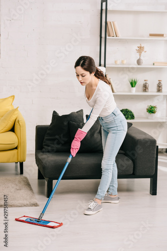 young housewife washing floor with mop near grey sofa