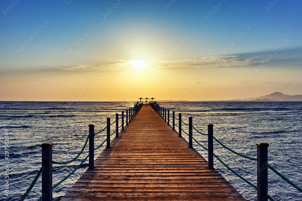 Bright and colorful sunrise over the sea and pier. Perspective view of a wooden pier on the sea at sunrise with rocky islands in the distance Bright and colorful sunrise over the sea and pier. Perspective view of a wooden pier on the sea at sunrise with rocky islands in the distance
