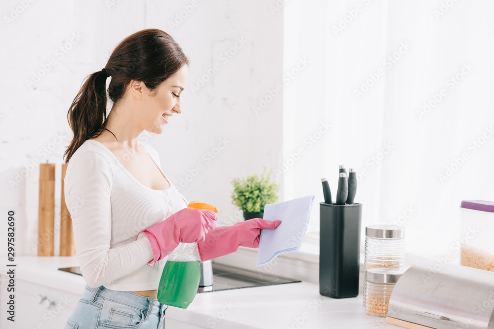 cheerful housewife spraying detergent from spray bottle on rag in kitchen cheerful housewife spraying detergent from spray bottle on rag in kitchen