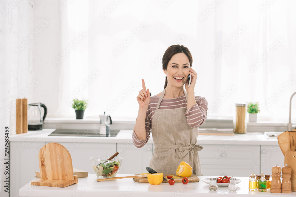 cheerful woman in apron showing idea sign while standing near kitchen table with vegetables and talking on smartphone cheerful woman in apron showing idea sign while standing near kitchen table with vegetables and talking on smartphone