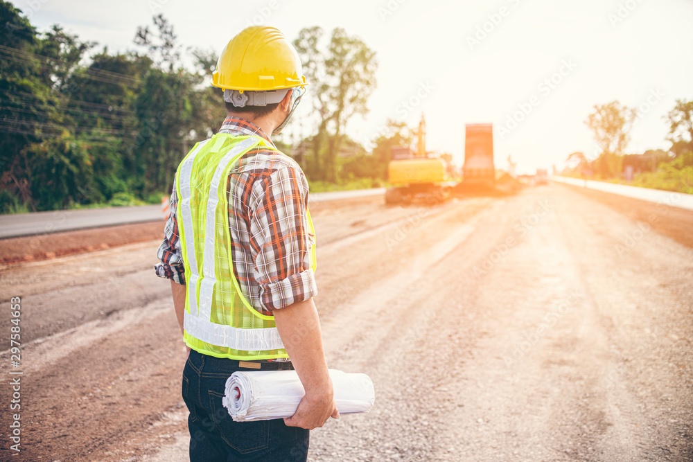 Construction workers or engineers wear reflective vests and carry helmets. And stood in front of a large machinery excavator during the construction of the highway