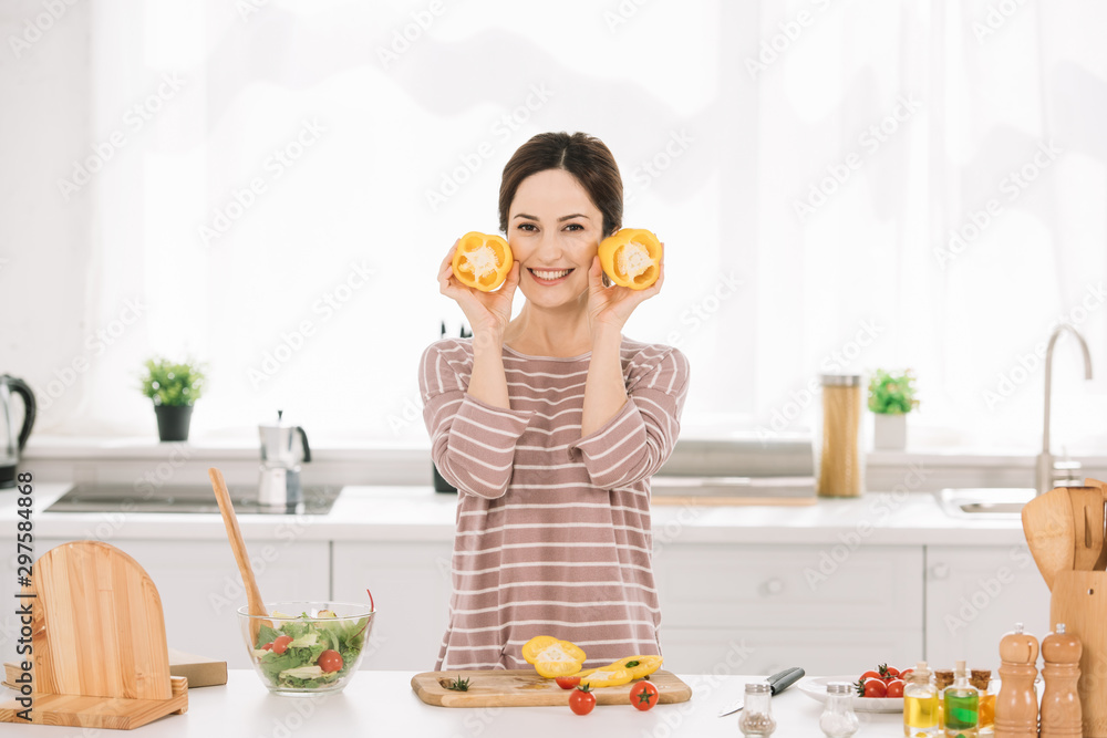 attractive young woman looking at camera while standing near kitchen table with fresh vegetables and holding cut bell pepper