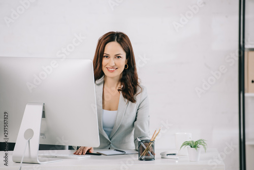attractive, smiling secretary looking at camera while sitting at workplace photo