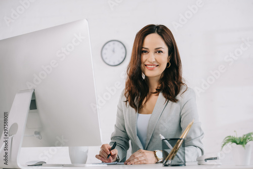beautiful, smiling secretary looking at camera while sitting at workplace near computer monitor