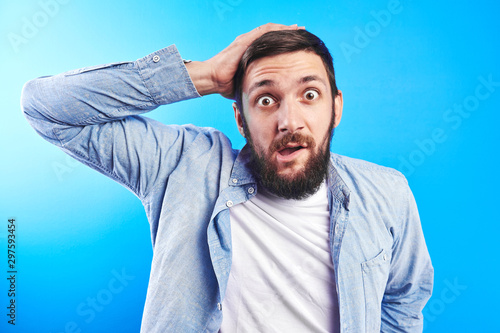 Shocked Caucasian bearded man with bulging eyes and open mouth in surprise clutched his head isolated in blue studio. Discounts, low prices, sale in store