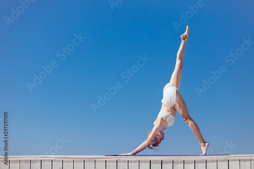 Sporty young woman doing stretching standing in One legged dolphin pose. Slim girl practicing yoga outdoor by the sea or ocean  blue sky  white wooden terrace. Calm  relax  healthy lifestyle concept