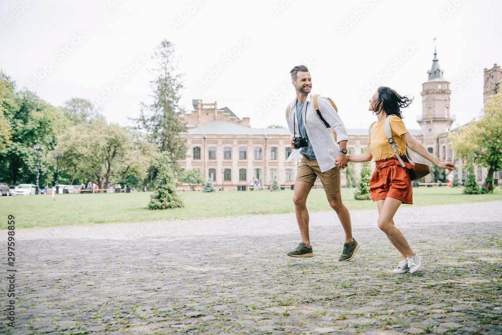 Obraz premium cheerful bearded man and woman holding hands while running near building