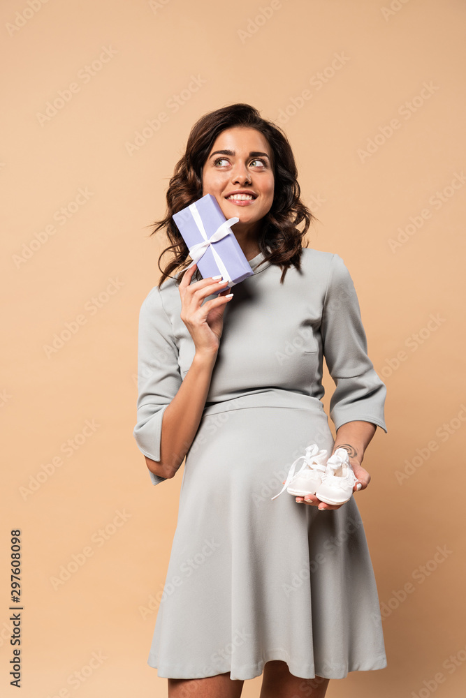 pregnant woman in grey dress holding gift and shoes on beige background pregnant woman in grey dress holding gift and shoes on beige background