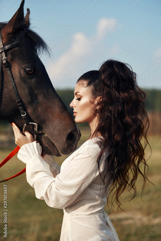 Obraz premium Girl in a long dress stands near a horse, a beautiful woman strokes a horse and holds the bridle in a field in autumn. Country life and fashion, noble steed