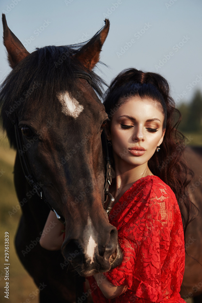 Fototapeta premium Girl in a long dress stands near a horse, a beautiful woman strokes a horse and holds the bridle in a field in autumn. Country life and fashion, noble steed
