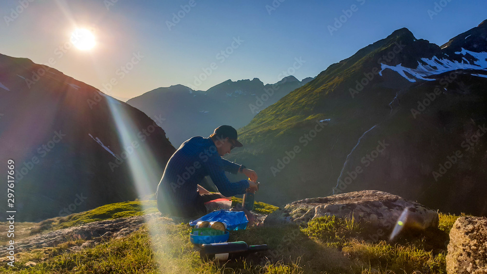 A young man cooking a dinner in the wilderness during the sunrise. He is sitting at the edge of a mountain, mixing the dish. He is surrounded by tall mountains. Alpine camping. Outdoor kitchen. - obrazy, fototapety, plakaty 