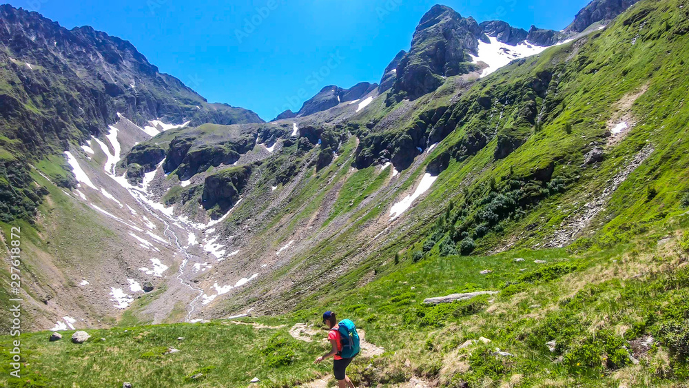 A young woman with a big backpack hikes down on a steep pathway between tall mountain peaks. Some of the slopes are covered with snow. In the back is another mountain range. Spring in alpine valleys.