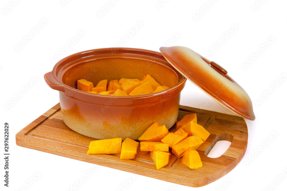 Fototapeta premium A small ceramic pan with an open lid filled with sliced pumpkin on a wooden cutting board on which pieces of pumpkin also lie. Isolated On white background.