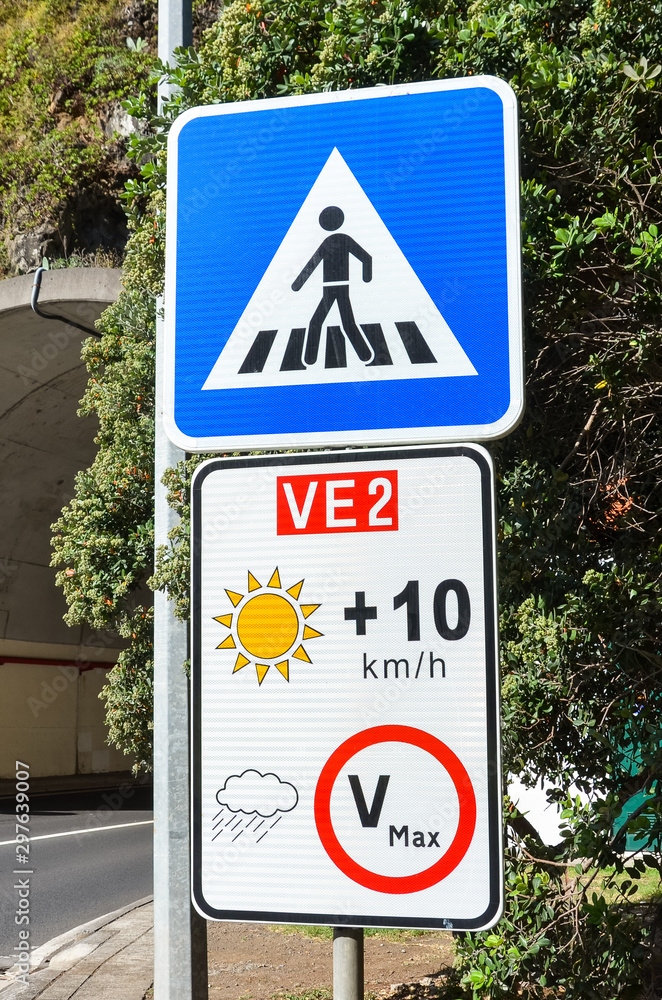 Road signs in Portuguese island Madeira. Blue zebra crossing sign. White speed limit sign. The speed restriction is dependent on weather, in case of clear weather the limit is plus 10 kmh than stated Road signs in Portuguese island Madeira. Blue zebra crossing sign. White speed limit sign. The speed restriction is dependent on weather, in case of clear weather the limit is plus 10 kmh than stated