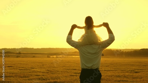 concept of happy childhood. dad with little daughter on their shoulders walk and watch beautiful sunset. family, father plays with his little child in park. concept of teamwork and growth in business