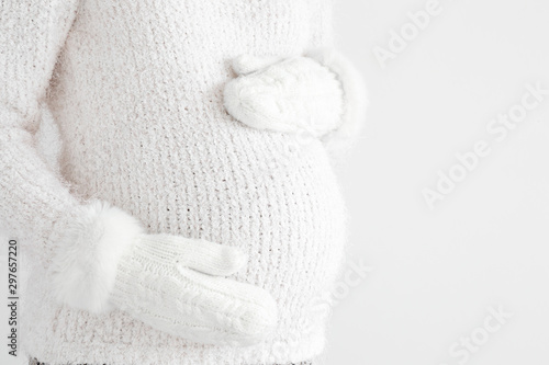 Pregnant woman in white fluffy sweater touching big belly with hands in mittens. Isolated on gray background. Warm clothes in cold weather in pregnancy time. Baby expectation. Side view. Closeup.