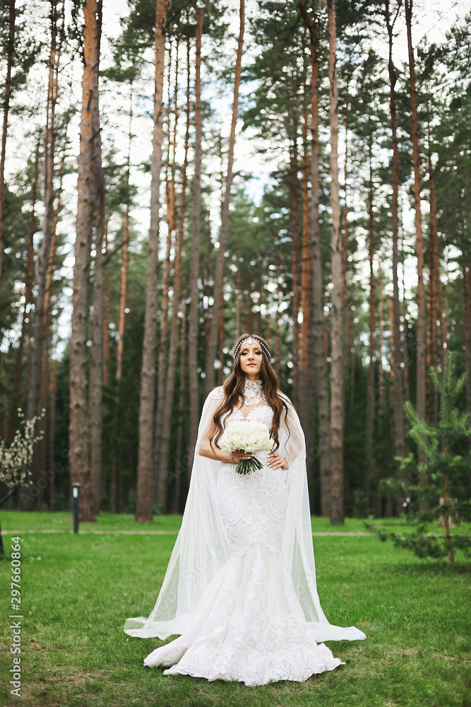 Young stylish bride with jewelry in her hairstyle wearing in lace wedding dress keeping a bouquet of flowers and posing in the forest. Beautiful woman in wedding outfit posing outdoors.