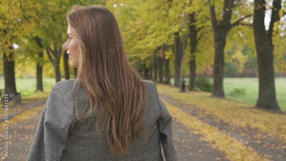 Confident Caucasian brunette girl looking around and walking away along the road. Nice smiling woman in checkered jacket strolling in the autumn park. Confident Caucasian brunette girl looking around and walking away along the road. Nice smiling woman in checkered jacket strolling in the autumn park.
