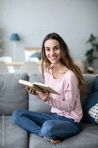 Woman with a book on sofa looking up at the camera with a smile.