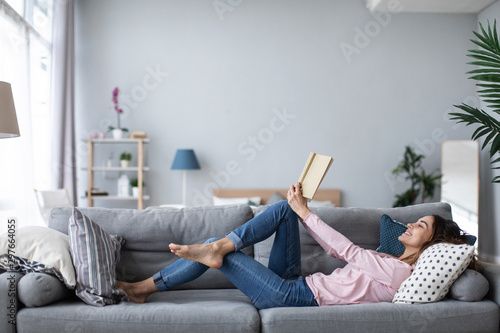 Young beautiful woman is reading a book while lying on a sofa in the living room.