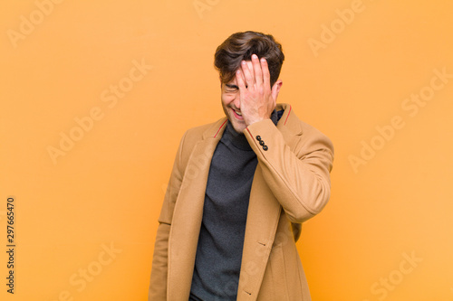 young handsome man laughing and slapping forehead like saying d‚Äôoh! I forgot or that was a stupid mistake against orange background