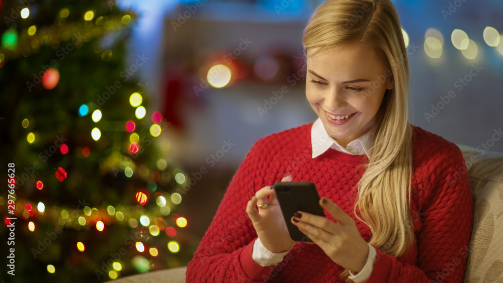 Beautiful Blonde Woman Sits on a Couch and Uses Smartphone. Christmas Tree and Room Decorated with Lights are in the Background. - obrazy, fototapety, plakaty 