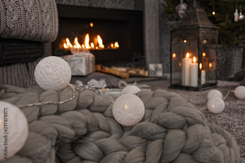 Beautiful winter composition in gray shades with a fireplace, cozy knitted blanket, candles, Christmas gifts and a garland.