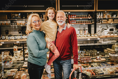 Happy grandmother and grandfather with granddaughter shopping together in grocery store or supermarket. Consumerism concept.