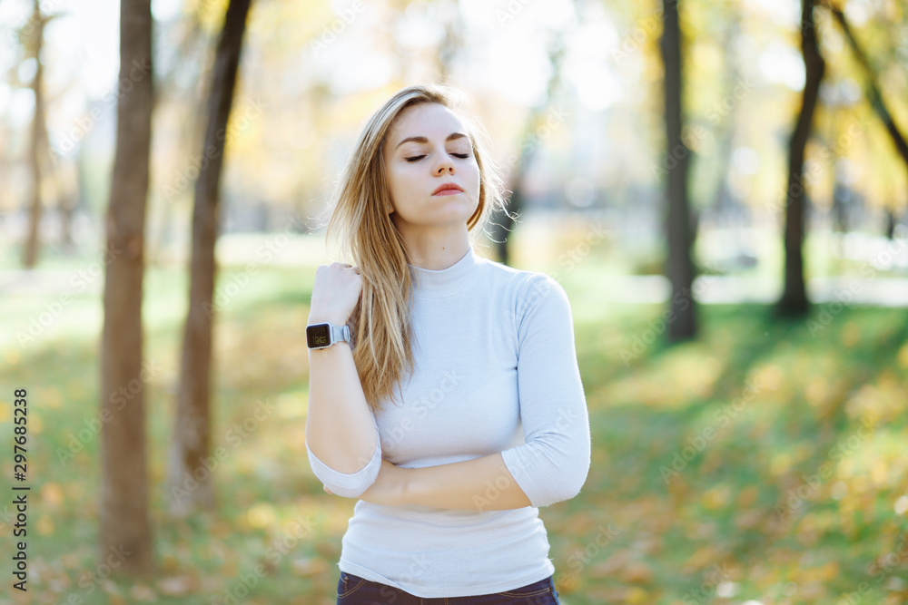 Happy young blonde woman in park on sunny autumn day. Cheerful beautiful girl in white sweater and smart wath on wrist. Retouched, vibrant colors, back lit, natural light. Happy young blonde woman in park on sunny autumn day. Cheerful beautiful girl in white sweater and smart wath on wrist. Retouched, vibrant colors, back lit, natural light.