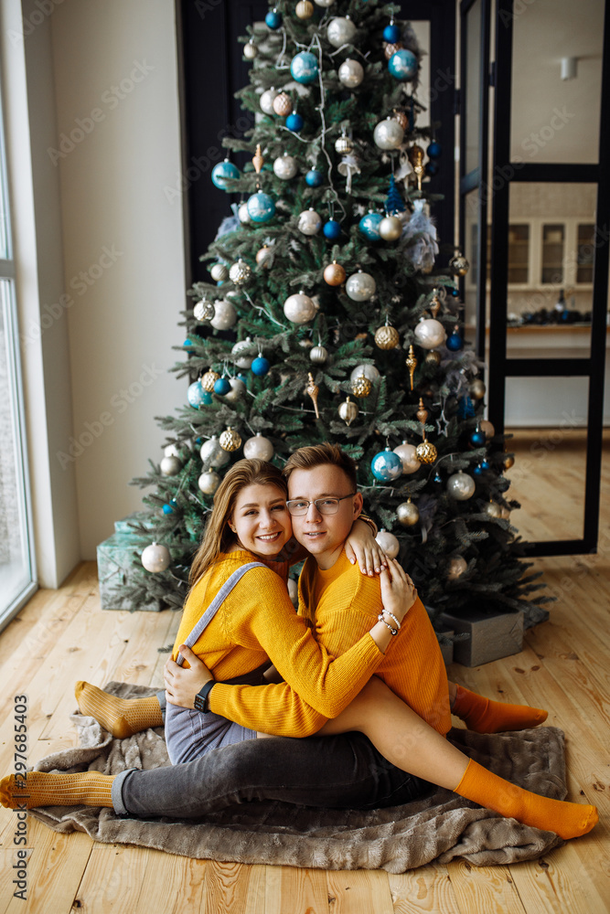 Happy couple, in stylish clothes, sitting on a rug and hugging each other near a Christmas tree. Christmas time. Christmas mood Happy couple, in stylish clothes, sitting on a rug and hugging each other near a Christmas tree. Christmas time. Christmas mood