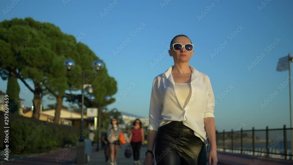 stylish bald girl in sunglasses and a white shirt walks the street against a blue sky and green trees stylish bald girl in sunglasses and a white shirt walks the street against a blue sky and green trees