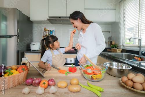 Portrait of beautiful Asian young woman and her daughter cooking salad for lunch and feeding tomato to her mother while making food, Family life love relationship, or home fun leisure activity concept photo