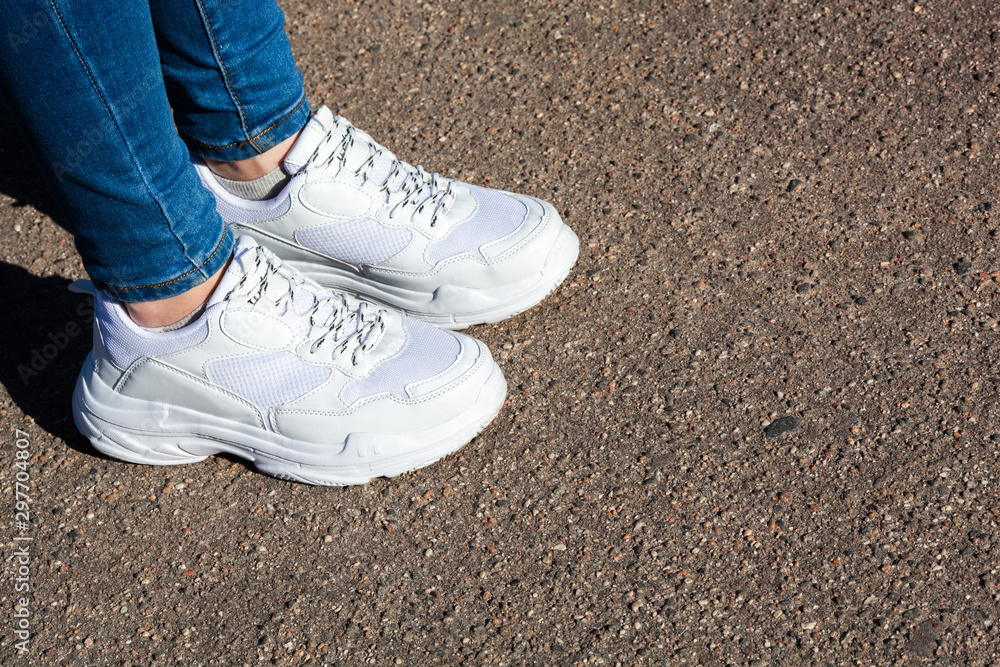 women's legs in white sports running shoes and jeans on the background of dry asphalt