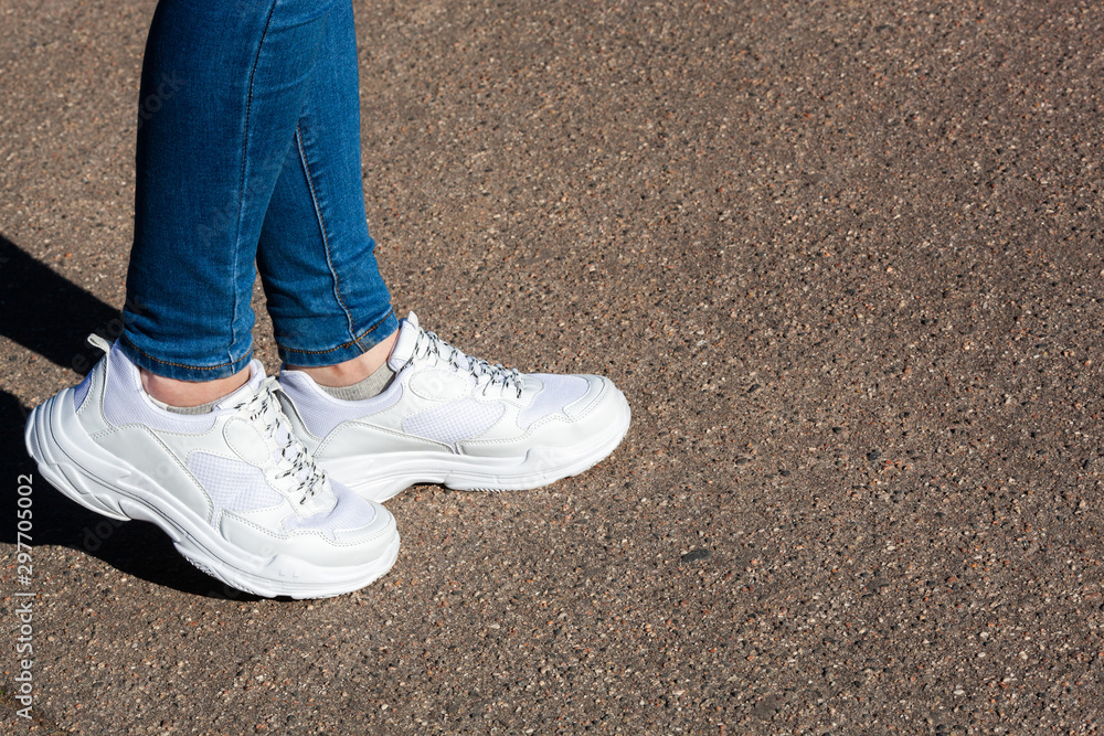 women's legs in white sports running shoes and jeans on the background of dry asphalt