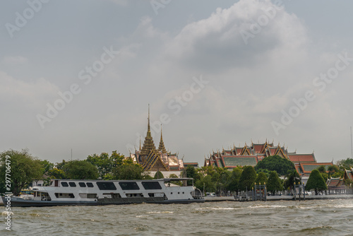 Bangkok city, Thailand - March 17, 2019: Chao Phraya River. Large white ferry boat passes in front of National Grand Palace under light blue cloudscape and green foliage in front. 