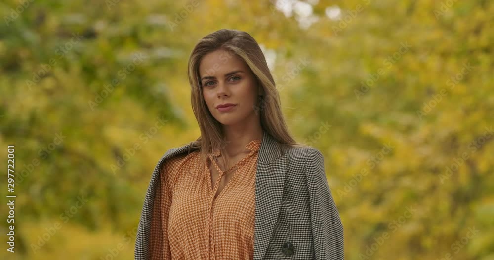 Smiling Caucasian woman with deep gaze standing on background of yellow leaves and looking at camera. Pretty woman in mustard dress and checkered jacket posing in the park. Cinema 4k footage ProRes HQ Smiling Caucasian woman with deep gaze standing on background of yellow leaves and looking at camera. Pretty woman in mustard dress and checkered jacket posing in the park. Cinema 4k footage ProRes HQ