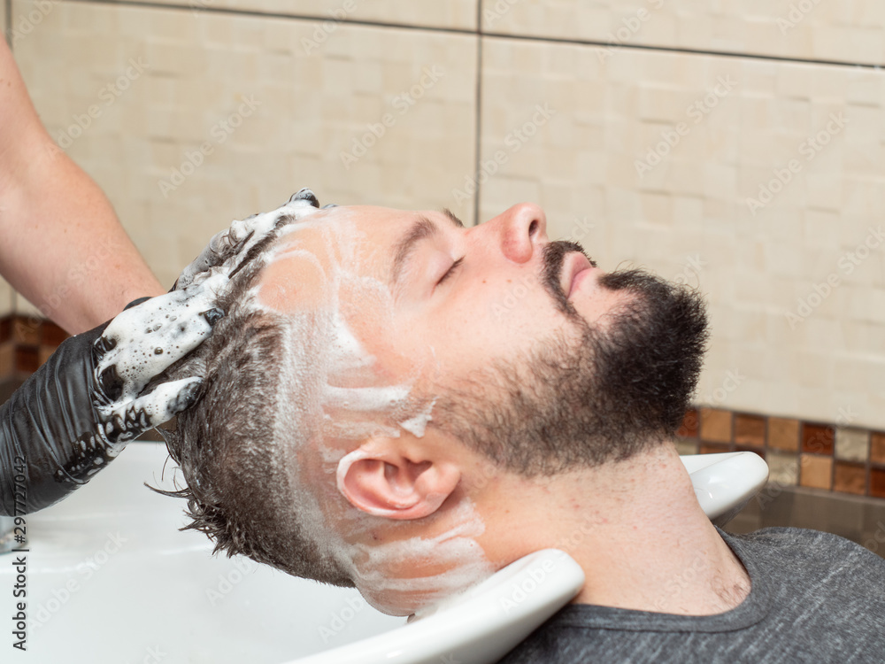 Obraz premium Stylist shampoo males hair in wash stand, close up view. Hairdressers hands in black rubber gloves. Male in barbershop. Selective soft focus. Blurred background