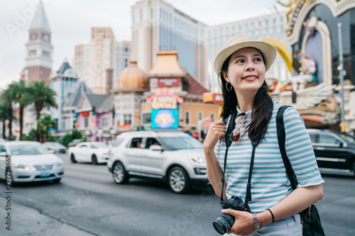 Happy girl travel in summer las vegas strip. smiling young woman backpacker holding vintage camera looking aside. female tourist stand next to busy road with cars and skyscrapers on background.
