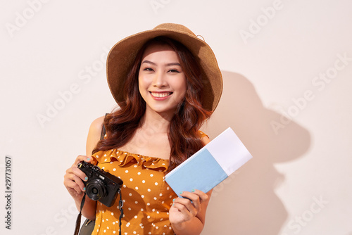 Portrait of a happy young woman in hat holding camera and showing passport while standing isolated over beige background