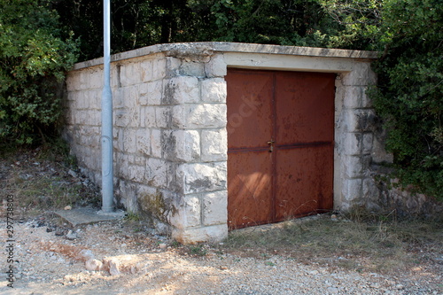 Detached small garage with rusted metal doors and stone walls next to gravel road surrounded with dense trees and vegetation on warm sunny summer day
