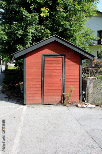 Locked dark red small wooden outdoor tool shed on edge of paved family house backyard next to tall tree on warm sunny summer day