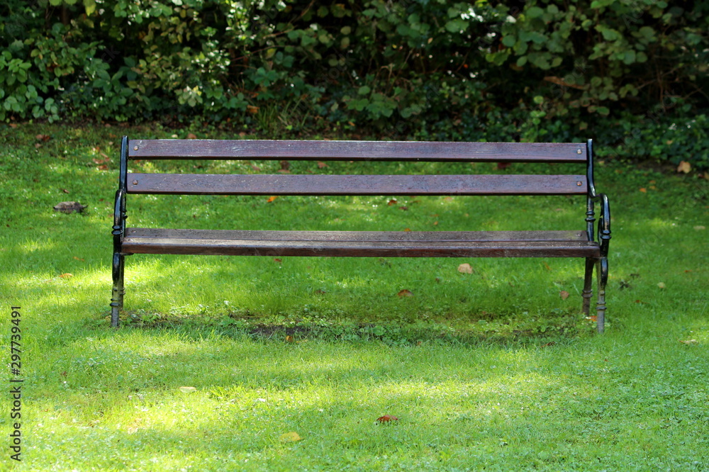 Old public park bench with wooden boards and baroque style wrought iron supports surrounded with fresh green grass and dense hedge in background on warm sunny summer day Old public park bench with wooden boards and baroque style wrought iron supports surrounded with fresh green grass and dense hedge in background on warm sunny summer day