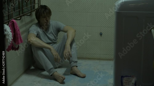 Full body shot of disheveled young man sitting on floor of dark creepy room