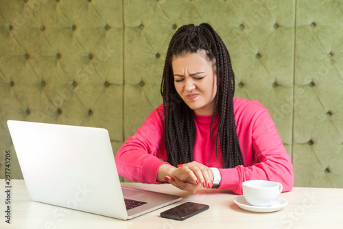 hand pain after too long working and typing owerwork on laptop. young woman in pink blouse is sitting in cafe and holding arm trying to feel the pulse with grimacing face. Indoor, health care photo