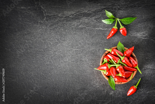 Hot pepper in wooden bowl on dark stone table. Chili red small peppers and green leaves on black background top view and copy space.