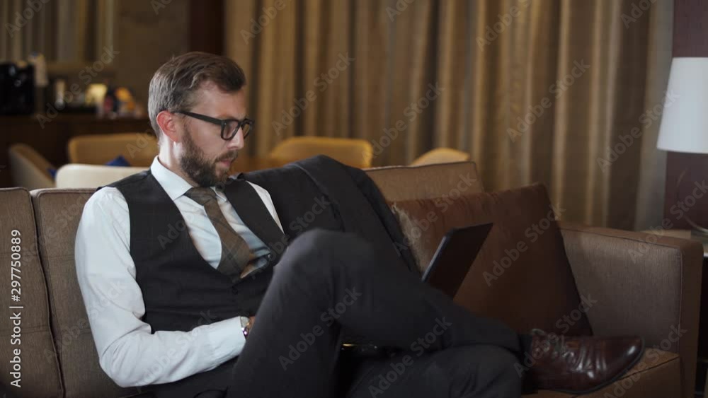 Young businessman in glasses working at a laptop, man in the suit sitting on the couch in the hotel, business trip. Young businessman in glasses working at a laptop, man in the suit sitting on the couch in the hotel, business trip.