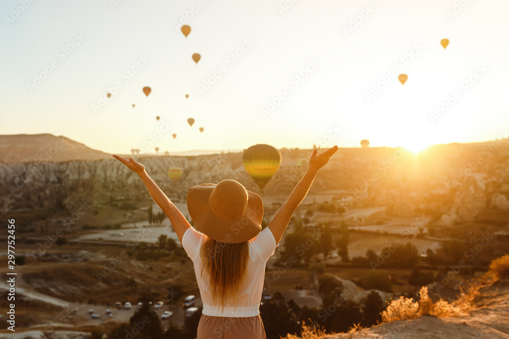 Beautiful young attractive girl in a hat stands on the mountain with flying air balloons on the background. Girl in the sunrise. View from the back. Famous tourist Turkish region Cappadocia. - obrazy, fototapety, plakaty 