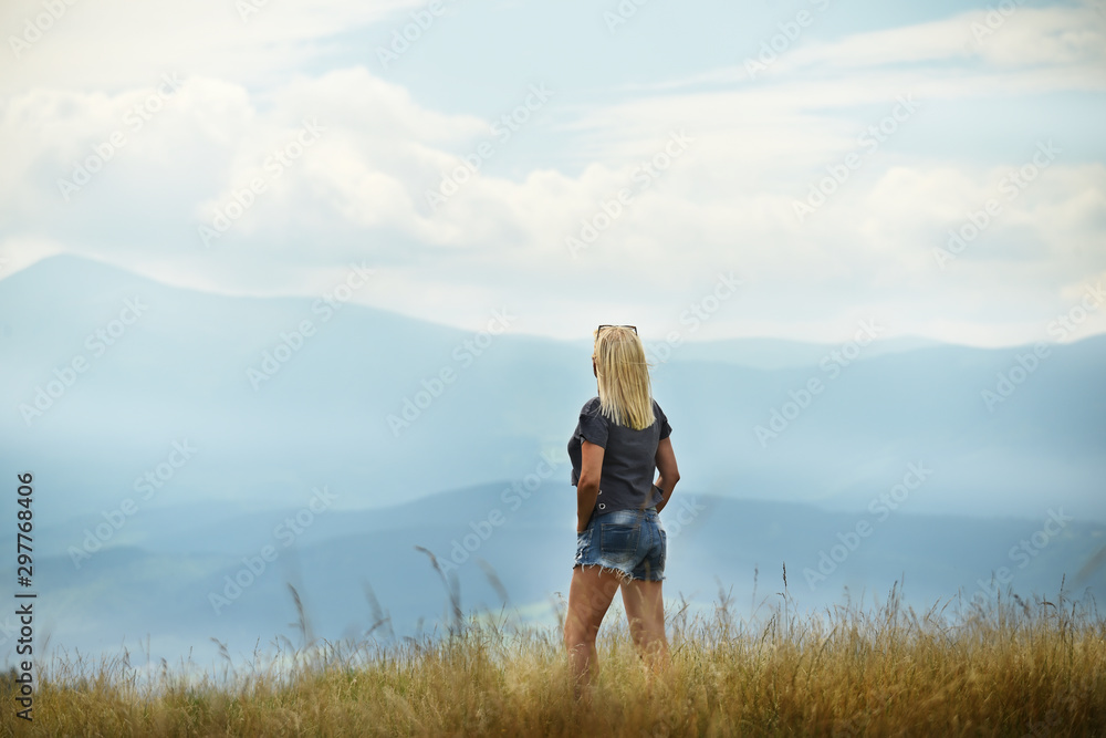 A girl standing with her back against the backdrop of a majestic view of mountains in the clouds. The atmosphere of travel and relaxed observation of nature.